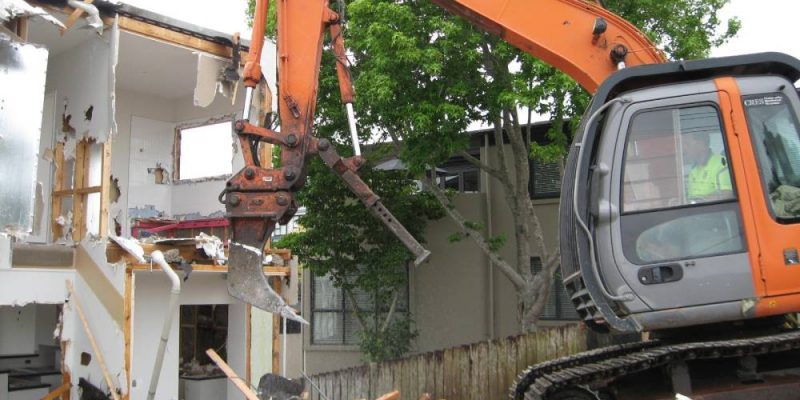 An orange excavator pulling down the wall of an residential building