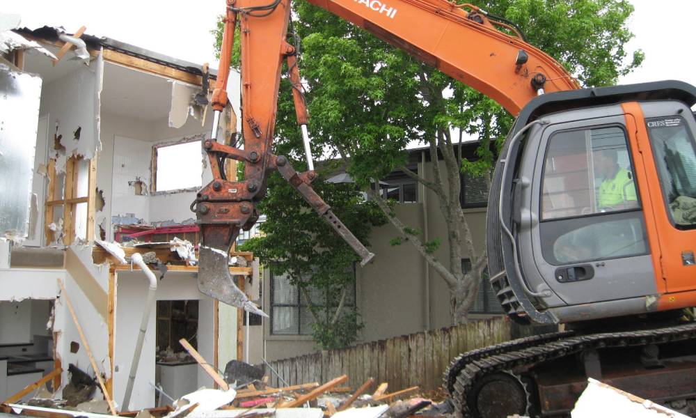 An orange excavator pulling down the wall of an residential building